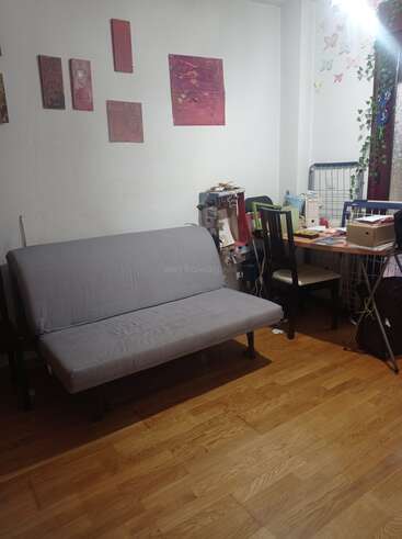 Una acogedora habitación con un futón gris, suelo de madera, una pequeña mesa de comedor, sillas, arte mural, papeles esparcidos y adornos de mariposas junto a una ventana con plantas.