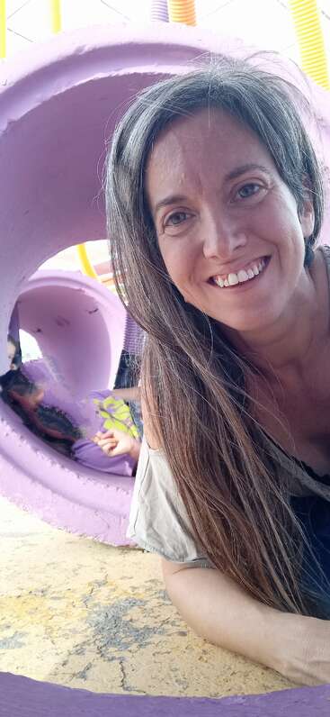 Una mujer con el pelo largo sonríe a la cámara, tumbada en el suelo cerca de unos tubos morados. Un niño juega dentro del tubo. Ambiente alegre, juguetón y luminoso.