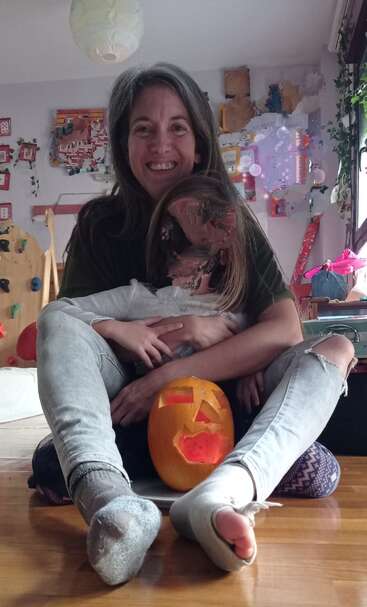 Una mujer sonriente abraza a un niño sentada en un suelo de madera. Entre los dos hay una calabaza tallada. La habitación es acogedora y está decorada de forma creativa.