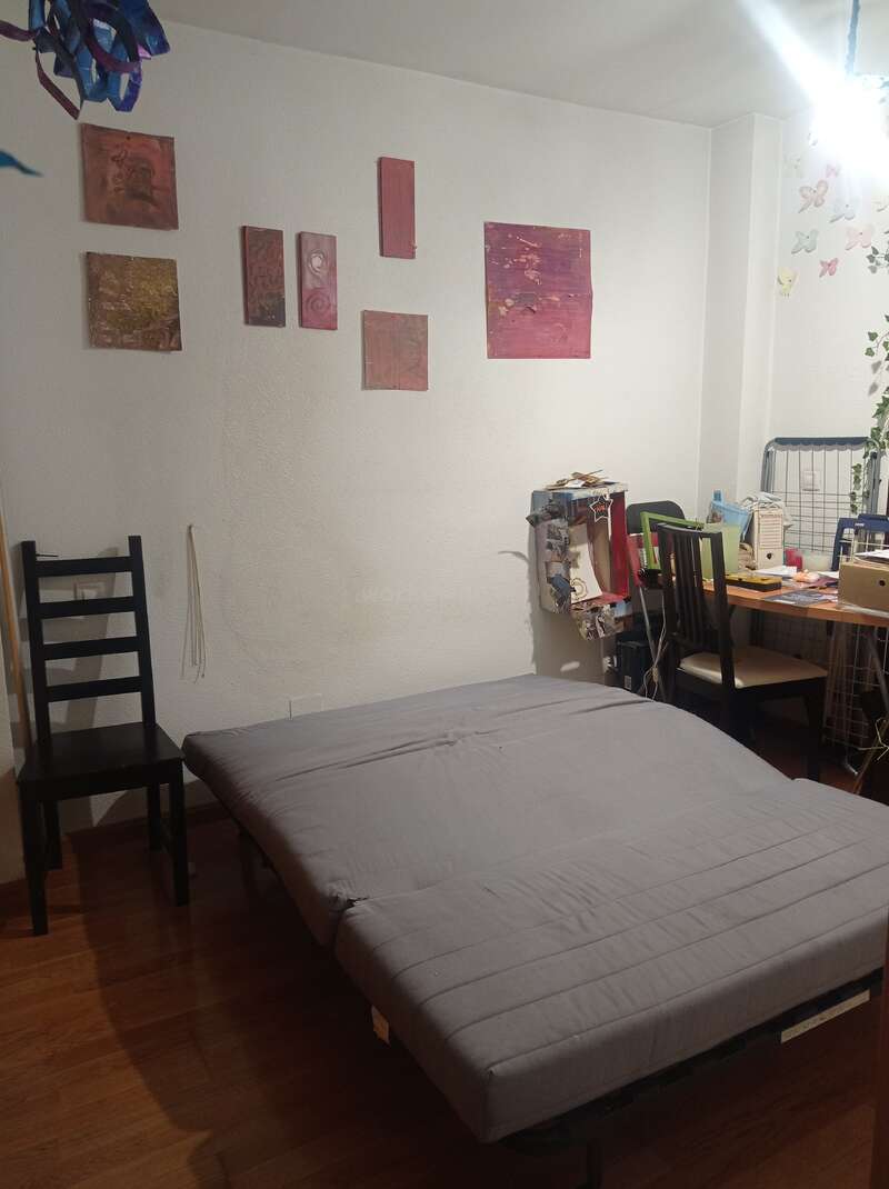 Una acogedora habitación con una cama gris, dos sillas negras, un escritorio repleto de objetos, arte en la pared, adornos de mariposas y suelo de madera iluminado por una luz brillante.