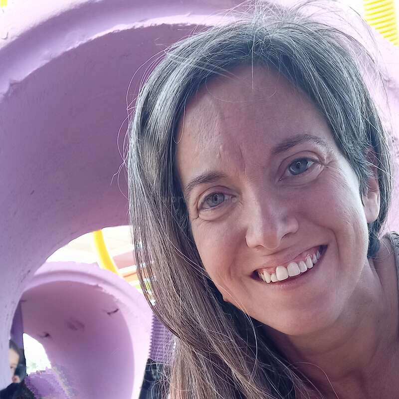 A woman with long hair smiles at the camera, lying on the ground near purple tubes. A child plays inside the tube. Bright, playful, joyful atmosphere.