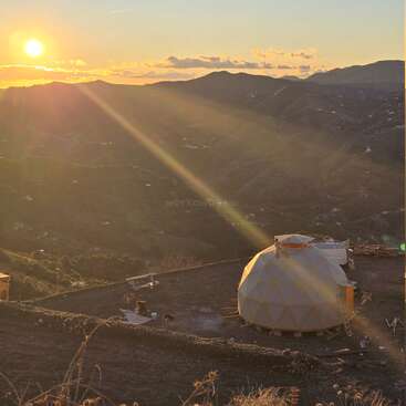 A golden sunset shines over rolling hills, illuminating a geodesic dome structure on a hillside. Soft sun rays spread across the serene, mountainous landscape.