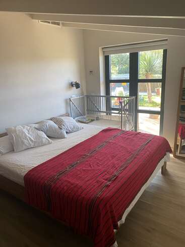 Ein helles Schlafzimmer mit einem großen Bett, einer rot gestreiften Decke, verstreuten Kissen, einem Kinderbett am Fenster, Holzboden und Blick auf einen Garten.