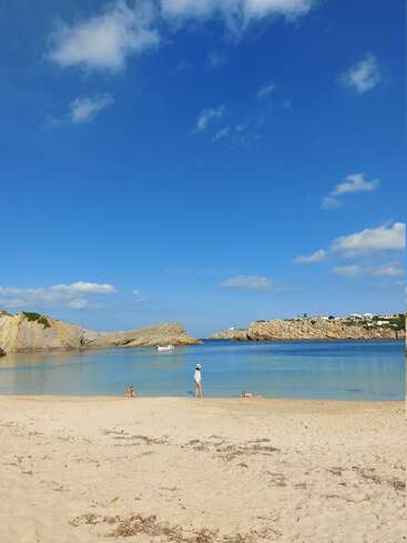 Ein ruhiger Sandstrand mit klarem, blauem Wasser, felsigen Klippen und strahlend blauem Himmel. Eine Person und zwei Hunde spazieren gemächlich am Ufer entlang. Eine entspannte Szene.