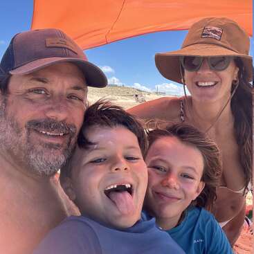 Eine glückliche vierköpfige Familie posiert gemeinsam unter einem orangefarbenen Sonnenschirm am Strand, lächelt und genießt den sonnigen Tag. Vorne stehen zwei Kinder.