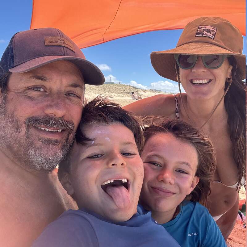 Eine glückliche vierköpfige Familie posiert gemeinsam unter einem orangefarbenen Sonnenschirm am Strand, lächelt und genießt den sonnigen Tag. Vorne stehen zwei Kinder.