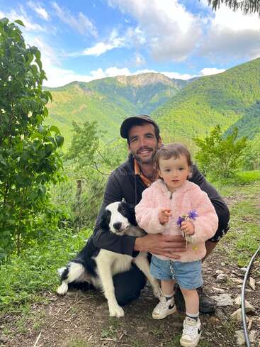 Un homme, un petit enfant tenant des fleurs et un chien noir et blanc posent ensemble en plein air, avec en arrière-plan des montagnes verdoyantes, des arbres et un ciel bleu.
