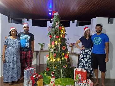 Four people wearing Santa hats stand beside a decorated Christmas tree made of green leaves, surrounded by gifts, smiling and celebrating Christmas indoors under warm lighting.