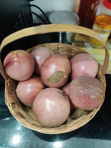 A wicker basket filled with several round, reddish-purple passion fruits sits on a kitchen counter, surrounded by spice jars and metal containers in the background.