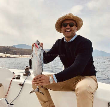 A man sits on a boat, holding a fish with a hook in its mouth, wearing a straw hat, sunglasses, and a black sweater.