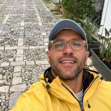 Ein Typ mit Brille, blauer Mütze und gelber Jacke lächelt für ein Selfie auf einer Kopfsteinpflasterstraße, die von Topfpflanzen und Grünpflanzen gesäumt ist.