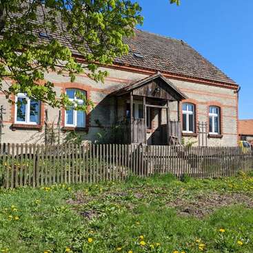 The image depicts a charming, rustic house with a wooden fence, surrounded by a lush green lawn dotted with yellow flowers, set against a clear blue sky.