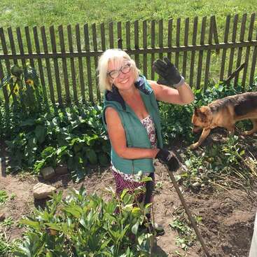 Eine lächelnde ältere Frau in Gartenkleidung winkt fröhlich, während sie in einem sonnigen Garten arbeitet. Ein Hund streift in der Nähe umher, und ein Holzzaun bildet den Hintergrund.
