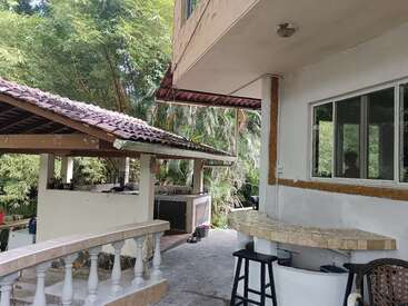 A shaded outdoor kitchen area beside a house, surrounded by lush green trees, features tiled roofs, a bar counter, stools, and a relaxing, tropical ambiance.