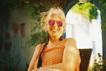 A smiling woman with blonde hair wearing pink sunglasses and an orange polka dot top sits outdoors in sunlight, surrounded by greenery and a relaxed atmosphere.