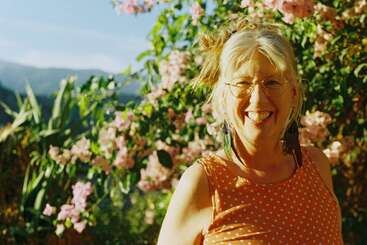 A cheerful woman with glasses and gray hair, wearing an orange polka-dot dress, smiles in a sunlit garden filled with pink flowers and greenery.