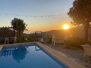 A tranquil sunset by a pool, surrounded by lush plants, string lights, cozy benches, and distant mountains. The warm light creates a peaceful, inviting atmosphere.