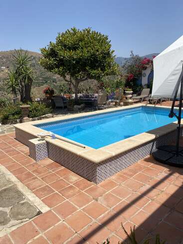 A serene outdoor pool surrounded by terracotta tiles, lush greenery, and comfortable seating overlooks scenic mountains under a clear blue sky on a sunny day.