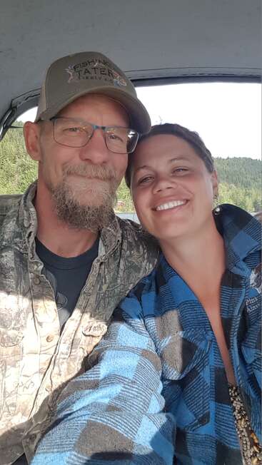 A smiling man and woman pose closely together, dressed in outdoor clothing. They appear happy and relaxed, possibly enjoying a day of fishing or nature. Trees behind.