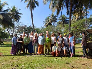 Un groupe diversifié de personnes se tient ensemble en plein air par une journée ensoleillée, entouré de palmiers et de verdure, souriant pour une photo de groupe dans un cadre décontracté.