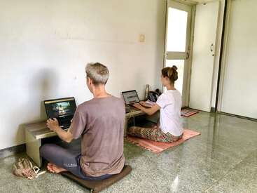 Deux personnes travaillent sur des ordinateurs portables, assises les jambes croisées sur des coussins et des tapis posés sur le sol, dans une pièce minimaliste et lumineuse, avec une petite table et une porte ouverte à proximité.
