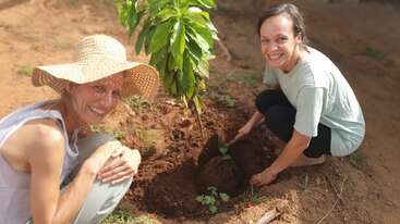 Deux femmes souriantes plantent un petit arbre dans un jardin. L'une porte un chapeau de soleil, l'autre une chemise légère. L'humeur est à la joie et à la collaboration.
