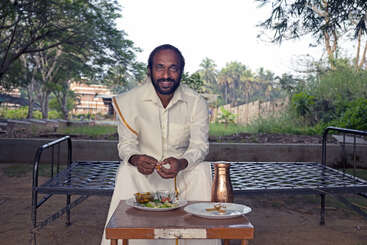 Un homme vêtu d'un costume traditionnel indien est assis en plein air sur un lit de camp, souriant, avec un repas et un gobelet en cuivre sur une petite table devant lui.
