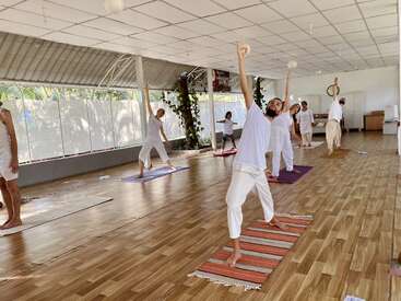 Un groupe de personnes pratiquant le yoga à l'intérieur sur des tapis, portant des vêtements blancs. Salle spacieuse avec sol en bois, lumière naturelle, verdure à l'extérieur et atmosphère paisible et concentrée.