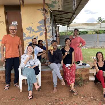 Un grupo de nueve personas, sonrientes y disfrutando de unas copas al aire libre junto a un edificio. Algunos están sentados, otros de pie, rodeados de plantas, ambiente relajado y alegre.