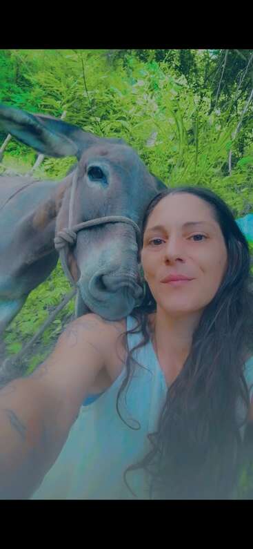Uma mulher de cabelos longos e escuros tira uma selfie com um burro simpático em um cenário ao ar livre, verdejante e exuberante. Os dois parecem relaxados e felizes juntos.