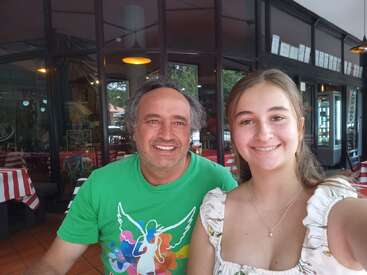 A man and a young woman are smiling, taking a selfie together at a restaurant with red and white tablecloths, glass windows, and a cheerful atmosphere.