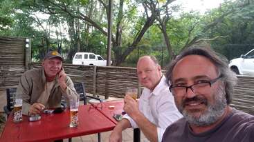 Three men sit around a red outdoor table, enjoying drinks and relaxing. Trees and parked white vehicles are visible in the background, creating a casual, friendly atmosphere.