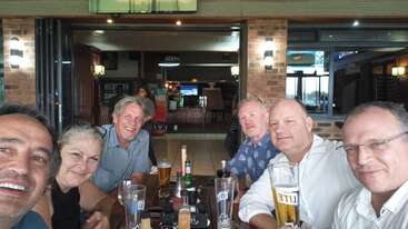 Six adults sit around a wooden table at a bar, smiling and enjoying drinks. The atmosphere is relaxed, with beverages and glasses visible on the table.