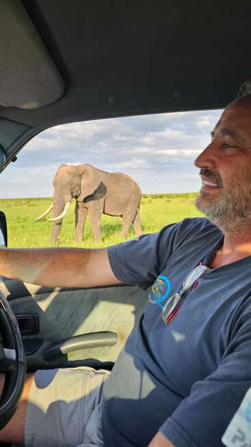 A man smiles while sitting inside a vehicle, with a large elephant walking in the grassy field outside under a partly cloudy sky, enjoying the safari.