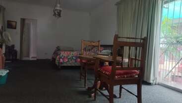 A cozy room with a floral bed, two wooden chairs, small table, large window with curtains, and a green trash bin. Light and airy ambiance.
