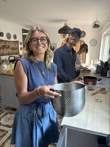 Three cheerful people are cooking together in a cozy kitchen. The woman in front smiles, holding a colander, while the others prepare ingredients in the background.