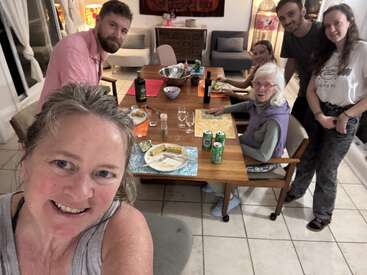 Six people are gathered around a dining table, smiling at the camera. Plates, drinks, and food are on the table. Warm, homey indoor atmosphere, friendly vibe.