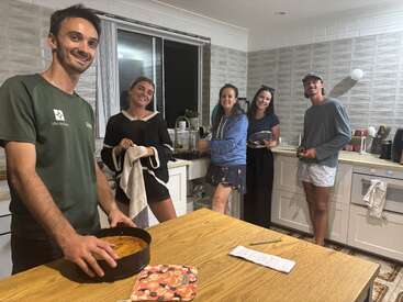 Five friends are smiling together in a cozy kitchen, preparing food and washing dishes. The atmosphere feels warm and relaxed, with everyone enjoying their time.