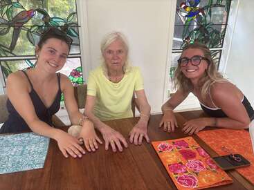 Three women, spanning generations, sit at a wooden table. They display their manicured nails, smiling together. Vibrant placemats and stained-glass windows featuring birds decorate the scene.