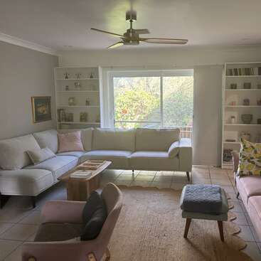 The image depicts a living room with white walls, a large window, and a light-colored couch, featuring a ceiling fan and bookshelves, with a serene garden view outside.