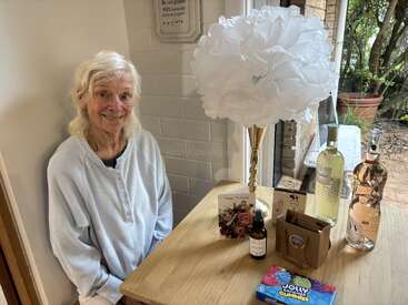 An elderly woman smiles beside a table decorated with birthday gifts, wine bottles, a large white floral arrangement, greeting cards, candies, and a window view.