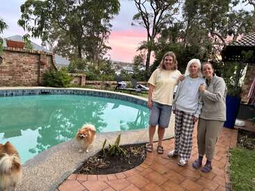 Three women stand together by a backyard pool at sunset, smiling warmly. Two small fluffy dogs are nearby. Trees, brick walls, and greenery surround them.