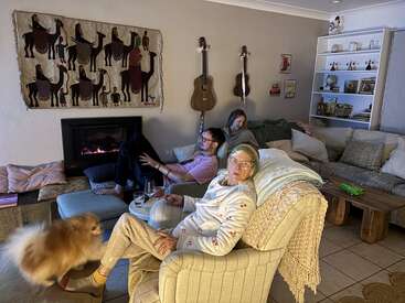 Três pessoas relaxam em uma aconchegante sala de estar, curtindo a lareira. Um cachorro fofinho está ali ao lado. Guitarras e quadros decoram a parede, e as estantes estão cheias de livros.