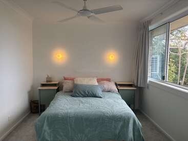 Um quarto aconchegante com uma cama de casal decorada com almofadas em tons pastéis e uma colcha azul-clara, duas mesinhas de cabeceira, luminárias de parede que criam um clima aconchegante e uma janela grande.