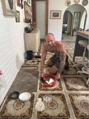 Um homem sem camisa e com tatuagens está ajoelhado em um piso de azulejos antigos, sorrindo enquanto limpa os detritos e os azulejos quebrados com uma pá de lixo numa cozinha aconchegante.