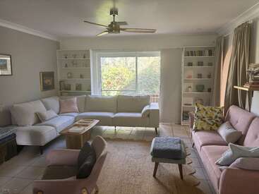 A imagem mostra uma sala de estar com paredes brancas, uma grande janela e um sofá de cor clara, com um ventilador de teto e estantes de livros, com uma vista serena para o jardim do lado de fora.