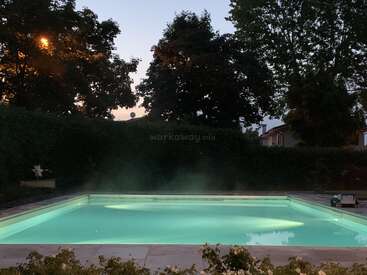 A serene, illuminated swimming pool glows at dusk, surrounded by lush greenery and silhouetted trees. Soft mist rises from the water, creating a tranquil, peaceful evening atmosphere.