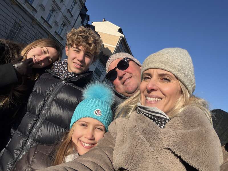 A family of five, bundled in winter clothing, smiles warmly for a selfie outside on a sunny day with blue skies and buildings in the background.