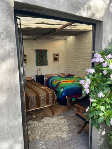 Una acogedora habitación rústica con dos camas, mantas de colores, tapices tejidos, paredes de ladrillo blanco, una alfombra de piel de oveja, luz natural y flores rosas en flor en el exterior.