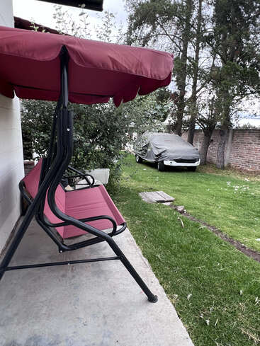 A red canopy swing sits on a concrete patio beside a white wall. In the background, a car covered in a tarp is on green grass.